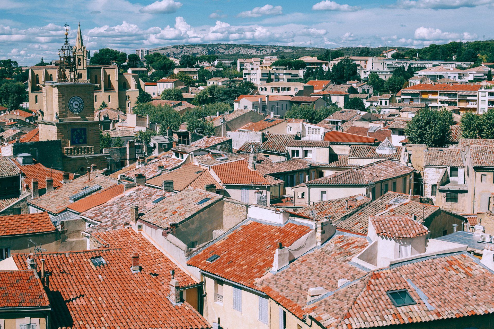 Vista de edificios con tejados de tejas rojizas, chimeneas y ventanas en las cubiertas, una torre con reloj y árboles bajo un cielo con nubes.